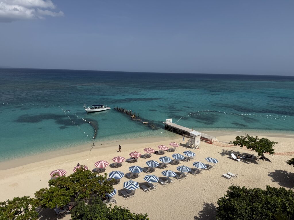 Aerial view of a white sand beach in Montego Bay, Jamaica, with turquoise Caribbean water, striped umbrellas, lounge chairs, and a boat anchored near the shoreline.